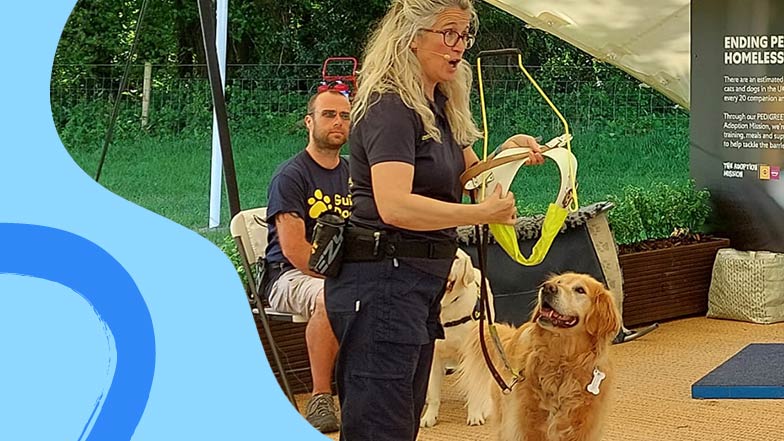 A member of the Guide Dogs demo team presenting while holding a harness with a golden retriever demonstration dog stood beside her