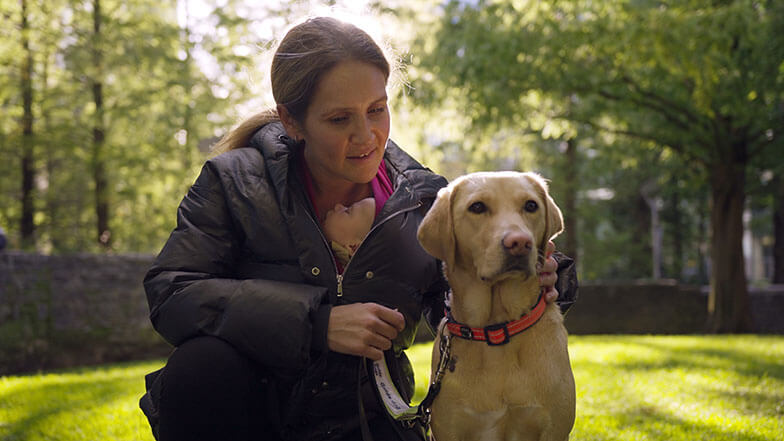 Guide dog owner Lorraine carrying her baby in a sling and crouched next to her yellow Labrador Retriever cross guide dog Theia