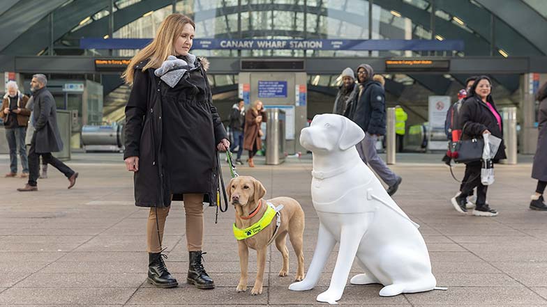 Guide dog owner Lorraine stood with guide dog Theia next to the blank white guide dog sculpture in front of Canary Wharf tube station