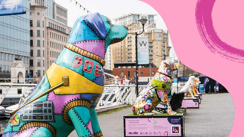 Image of a guide dog sculpture decorated as a robot situated in West India Quay. Three other decorated sculptures are shown lined up further down the path.