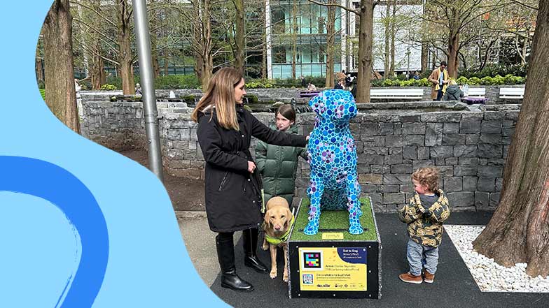 Image of a guide dog owner with her guide dog and two young children looking at a guide dog sculpture decorated with blue and purple spots.