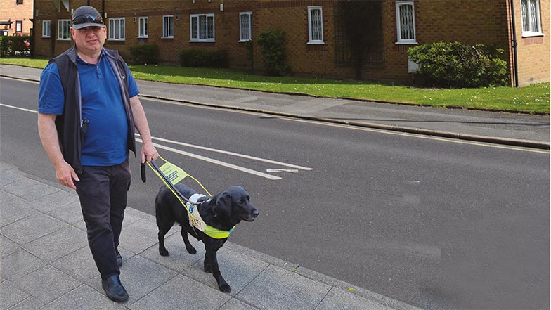 man and his guide dog in harness walking along a pavement