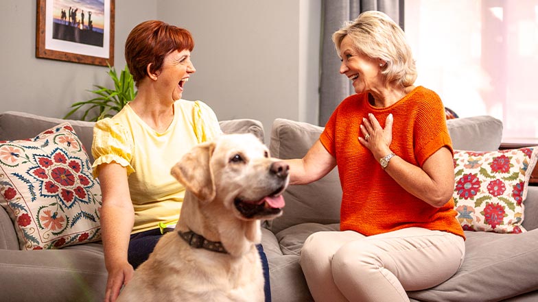 Two women looking excited sitting on a sofa, golden labrador in front of them 