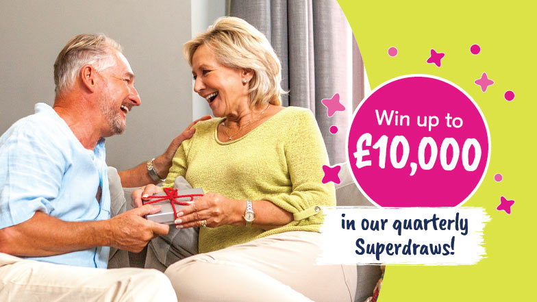 A man and woman sit together holding a present, the Superdraw prize of £10,000 is displayed.
