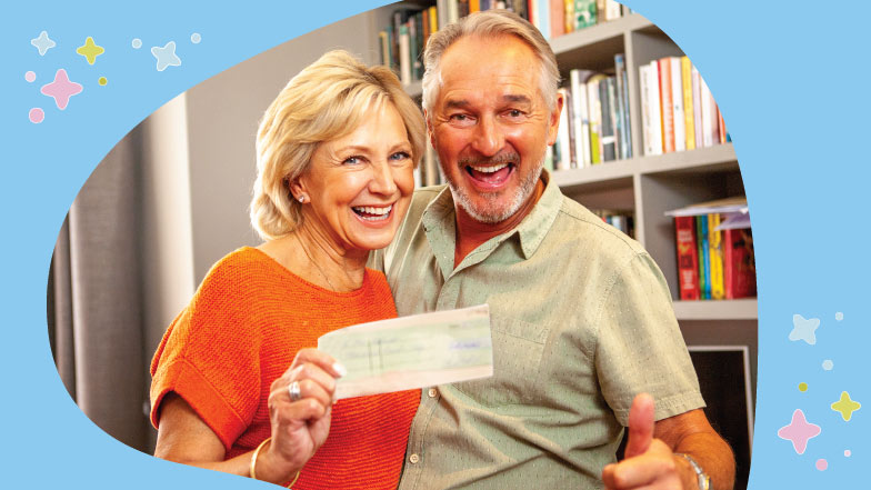 A couple stand together smiling and looking at camera while holding a cheque
