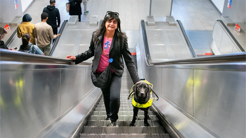 A woman and her black Labrador guide dog on an escalator in a shopping centre.