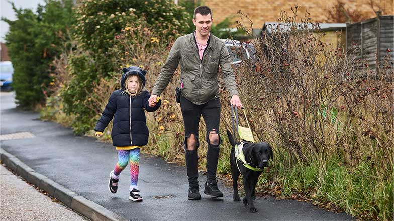 Guide dog owner Steve and his daughter walking with guide dog Wilf along a wet pavement.