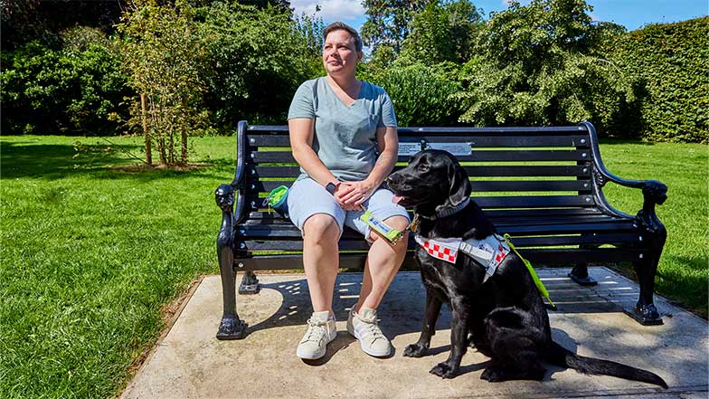 Guide dog owner Claire siting on a bench with black Labrador Jacqui in a park.