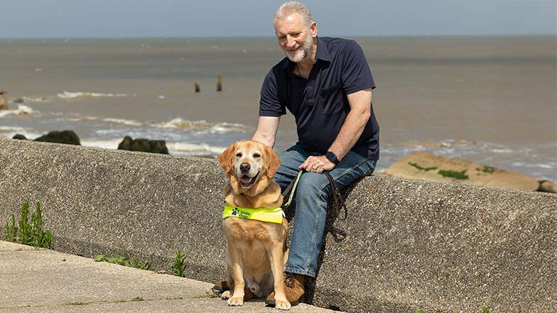 Guide dog owner Simon sitting on a sea wall with guide dog Mayne at his feet.