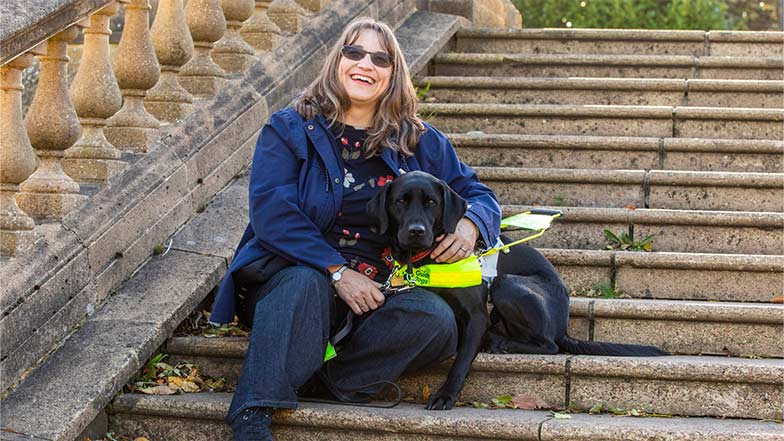 Guide dog owner Lesley and black Labrador Kide sitting together on outside steps.