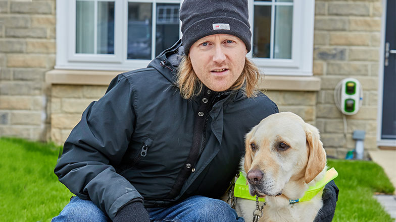 Chris and guide dog Jaeger in harness sitting together in front of a house