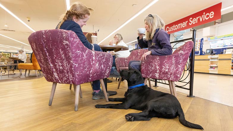 Guide Dog Trainer Victoria and Guide Dog Mobility Specialist Nicola sitting at a table with Sage laying at their feet. Guide Dog Trainer Victoria and Guide Dog Mobility Specialist Nicola sitting at a table with Sage laying at their feet.
