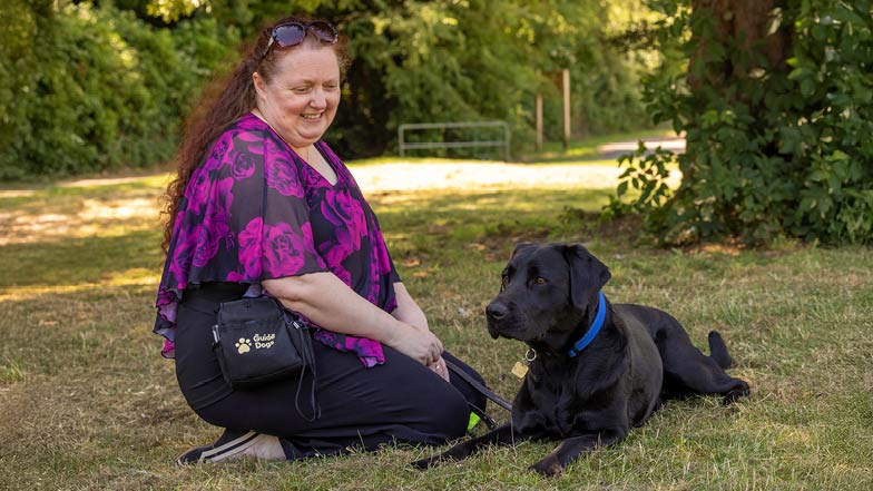 Sage lying on the grass next to smiling Guide Dog Owner Leonie who's sitting next to her. Sage lying on the grass next to smiling Guide Dog Owner Leonie who's sitting next to her.