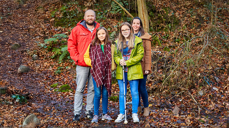 Caitlin, who has a vision impairment, stands with a white cane beside her family during a walk in the woods.