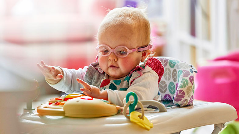 Margot, a baby with a vision impairment, plays on a baby walker. She is wearing glasses and appears engaged and curious.