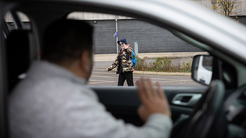A picture of a dad in car waving goodbye to his son who is going into school