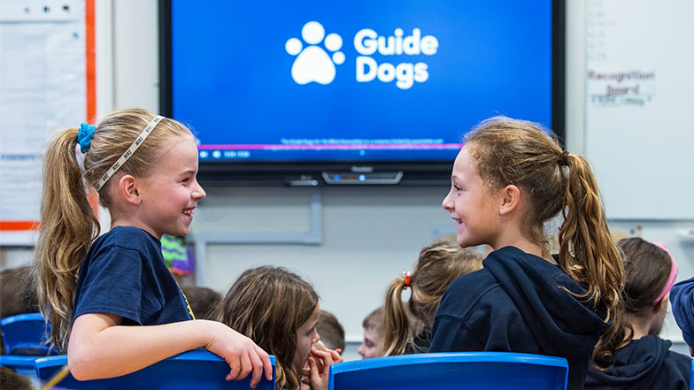 Two school girls smiling to each other in the classroom. The screen behind them shows the Guide Dogs logo.