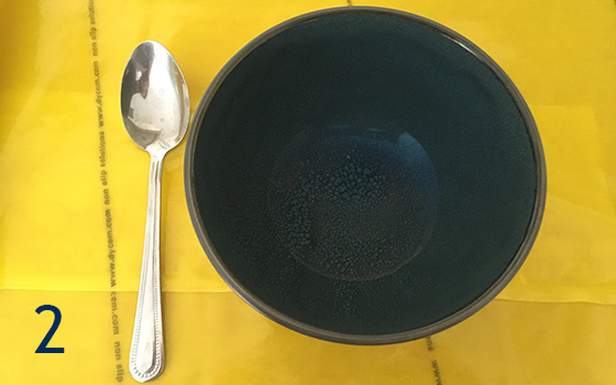 Image labelled with the number two. Shows a dark blue bowl and chrome spoon on a tray covered with yellow Dycem mat to highlight the contrast.