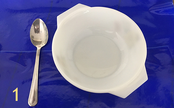 Image is labelled with the number one. Shows a white bowl and chrome spoon on a tray covered with a blue Dycem mat to highlight the contrast