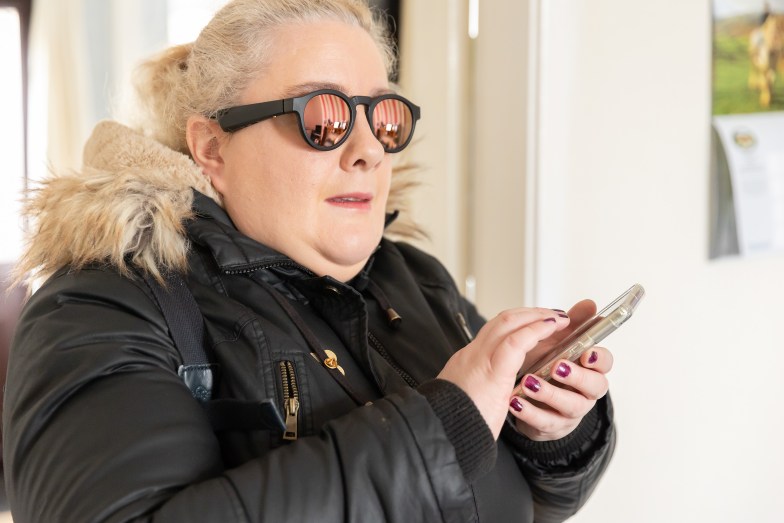 Marie, a woman with sight loss, stands indoors holding her phone as she uses an app.