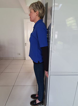 Picture of a Guide Dogs staff member standing against a wall with their, heels, bottom, back and shoulders all placed against the solid surface. 