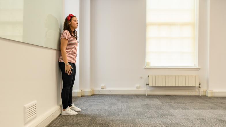 A woman stands against a wall in a meeting room, demonstrating the squaring off technique