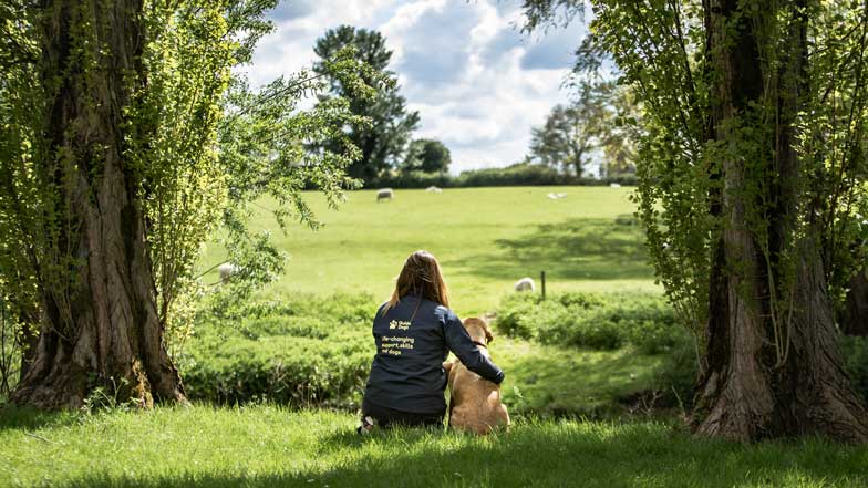 Guide dog trainer sat with their arm around a dog in training in green open fields
