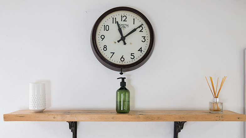 Image of a large black-rimmed clock on a white wall above a shelf.