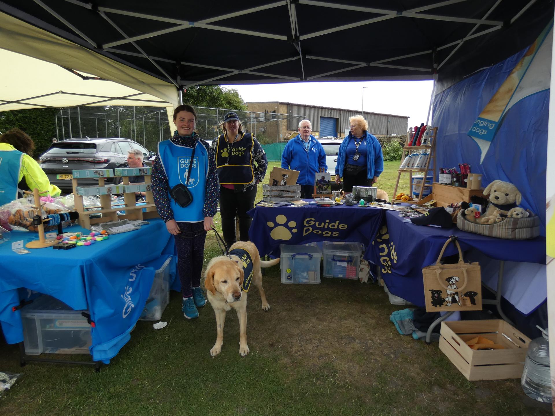 Members of the Airedale and Wharfedale fundraising group, including a yellow Labrador in a Guide Dogs training jacket, stand at a Guide Dogs stall