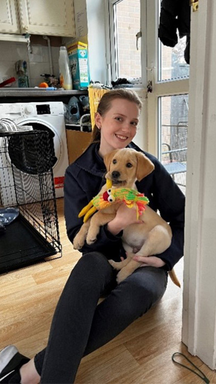 Puppy Development Advisor, Chloe Southby, who joined us this year sits on the floor, smiling at the camera with a yellow puppy in her lap. The puppy is holding a toy in its mouth