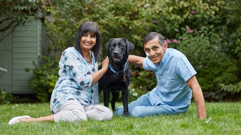 A man and woman are sitting on the lawn of their garden with a black puppy between them