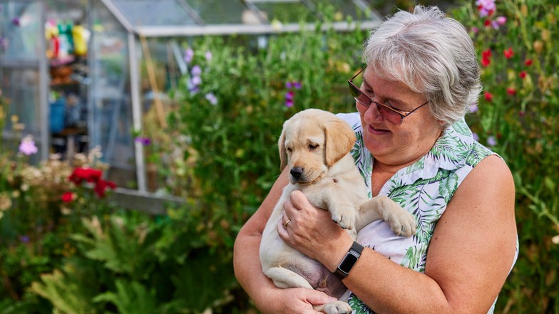 A puppy breeder is in her garden and is holding a puppy