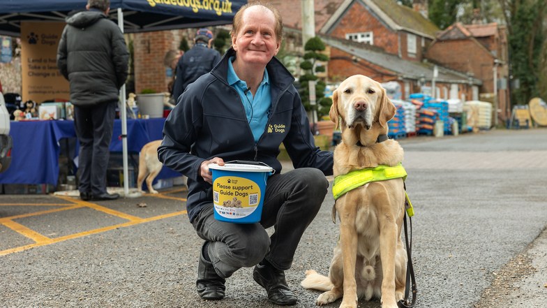 A volunteer fundraiser is crouched down next to a guide dog outside a fundraising stall
