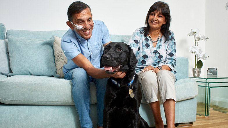 Two volunteer fosterers sit on a sofa with a black labrador guide dog before them