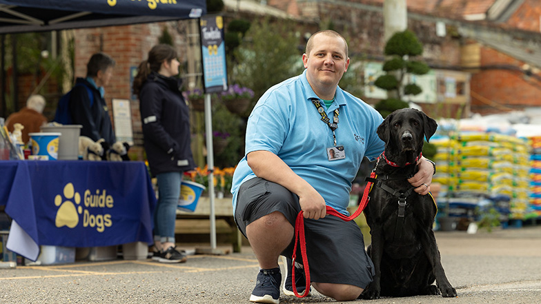Volunteer fundraiser smiles whilst knealing next to a guide dog