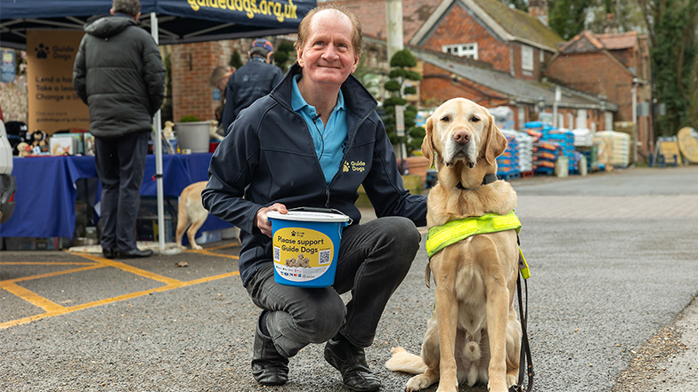 Volunteer fundraiser with a collection bucket sits with a guide dog