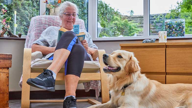 A volunteer Fosterer sat in a chair reading a book with a Golden Retriever sat on the floor beside her. 