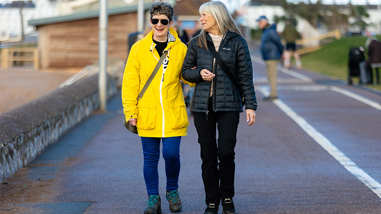 A woman with sight loss is being guided along a road by the beachfront by a female volunteer sighted guide