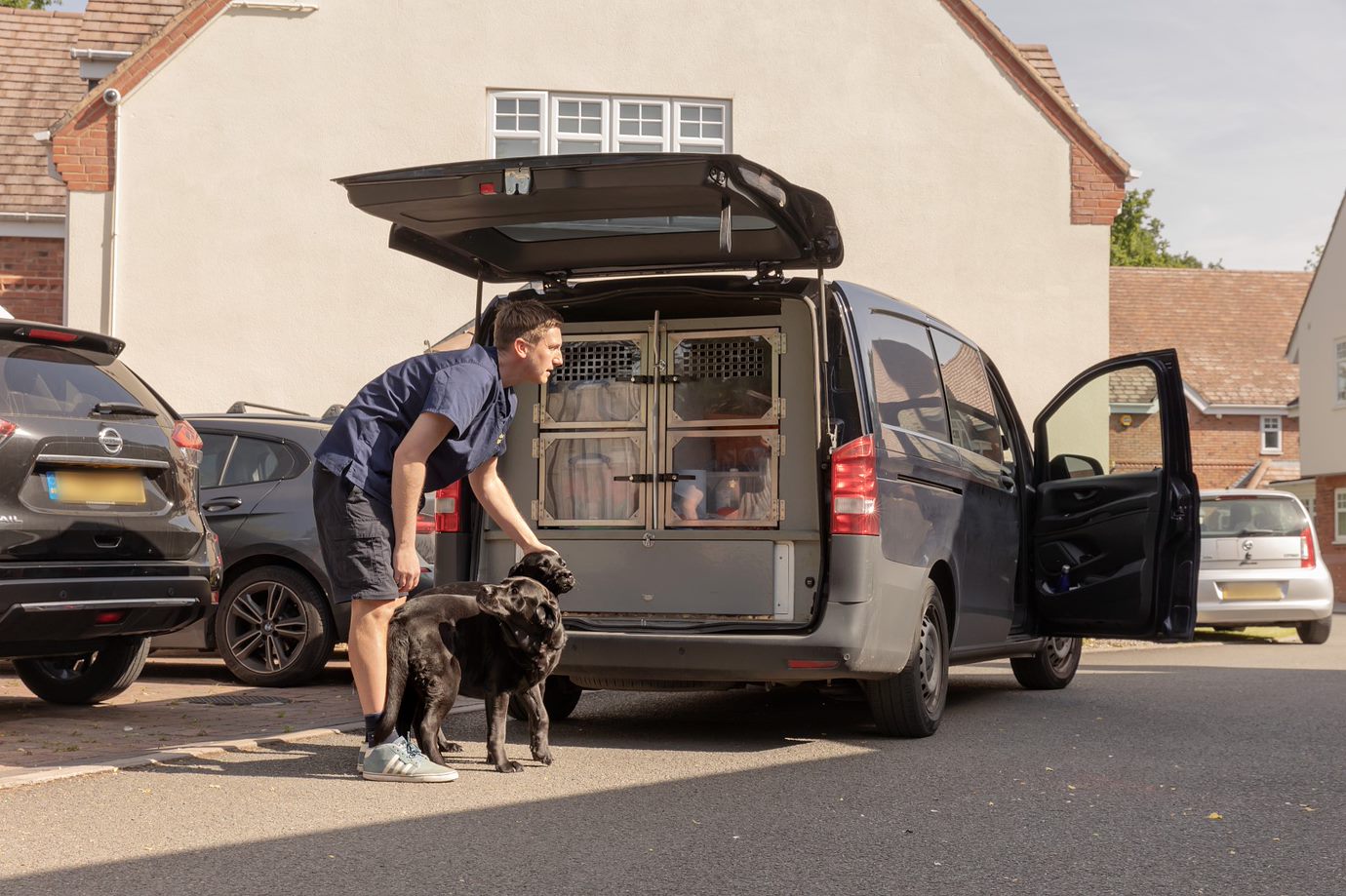 A mans stands before a van equipped to carry dogs, with two black labradors stood by the boot