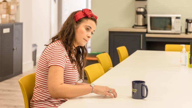 A woman sits at a table and makes circular motions to find an object. A mug sits on the table in front of her.