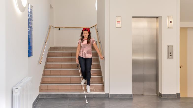A woman in a striped shirt and jeans walks down some stairs with her long cane