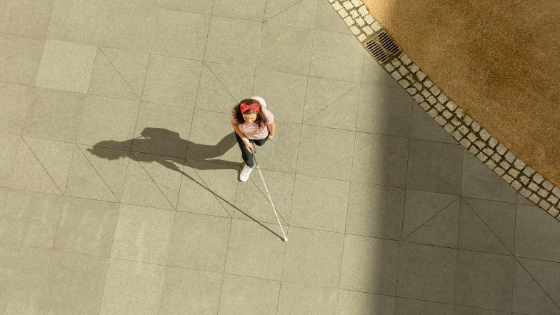 This aerial shot shows a long cane user walking across a sunny courtyard using the touch technique